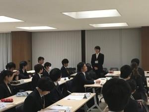 1名の女性が自分の席の前に立って話をしており、長机の席に座っている沢山の参加者が女性の方に体を向けている写真