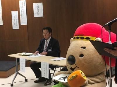 席に着き、机上の資料を見ている田渕教育長と池田市のキャラクター(ふくまるくん)の写真