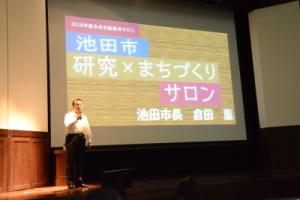 ステージの上の池田市研究×まちづくりサロンと映し出されたプロジェクタースクリーンの横に男性が立って話をしている写真