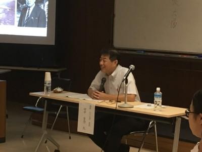 田中先生が両手でマイクを持ち笑顔で席に座っている写真