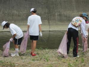 白い帽子を被り白いシャツを着た女子生徒たちが川岸のゴミ拾いをしている写真
