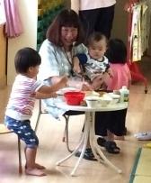 園長先生が赤ちゃんを膝に乗せ椅子に座り、男の子と一緒にままごと遊びをしている写真