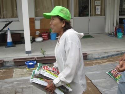 緑の帽子を被った女性が花や野菜などに使う肥料の入った袋を2袋持って歩いている写真