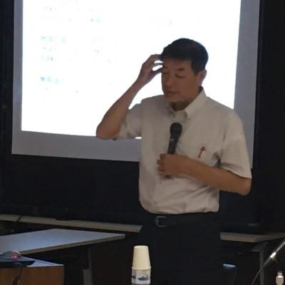 スクリーンの前で田中先生がマイクを持って話をしている写真
